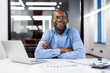 Obraz premium Businessman wearing glasses smiling confidently in office environment. He sits at desk with laptop and documents, exuding positivity and professionalism.