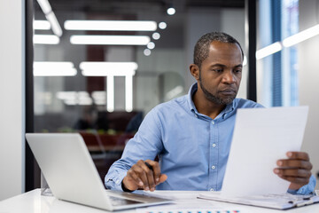 Businessman intensely examines documents at office desk with laptop open. Professional setting suggests roles like manager, analyst, or executive engaged in detailed work.