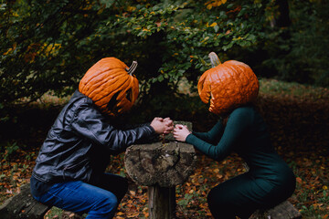 A couple, with pumpkins on their heads, sitting behind a wooden table in an autumn forest....