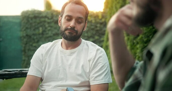 Over the shoulder a brunette guy with a beard in a white T-shirt listens attentively to his interlocutor friend and clinks beer with him while having fun in the backyard in the summer