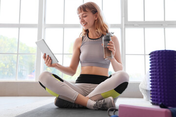 Young sporty woman with tablet and bottle of water resting after training at home