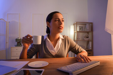 Tired young female programmer with cup of coffee working in office at night