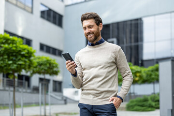 Businessman smiling while reading phone outdoors in urban setting. Professional in casual attire enjoying moment during break. Emphasizes career success, technology use, happiness, relaxation.