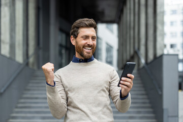 Smiling man with beard holding phone celebrating positive news or achievement. Expressing joy and...