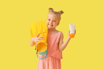 Cute little girl with sunscreen cream and flippers on yellow background