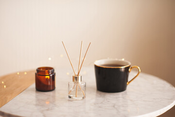 Home liquid perfume in glass bottle with bamboo sticks with burning candle with cup of tea on marble coffee table in living room close up. Cozy atmosphere.