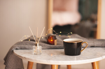 Home liquid perfume in glass bottle with bamboo sticks with cup of tea and scent candle on marble coffee table in living room close up. Cozy atmosphere.