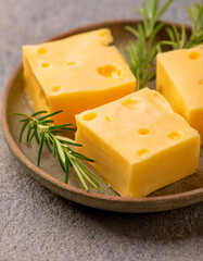 Creamy yellow cheese blocks on ceramic plate with fresh rosemary on stone table. Tasty food