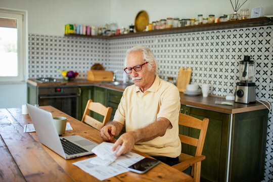 Elderly man managing bills and finances on laptop in kitchen