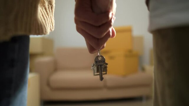 Close up shot hands of couple homeowner holding the house key and walking through inside the room with boxes and furniture background after moving to new house, relocating apartment concept.
