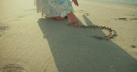 Close-up a girl in a white dress draws a heart on the wet sand on the beach during her morning walk