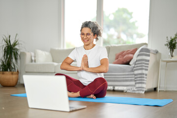 Happy healthy middle aged woman learning doing yoga at home with laptop. Serene mindful mature...
