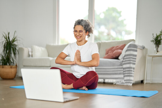 Happy healthy middle aged woman learning doing yoga at home with laptop. Serene mindful mature older lady in her 50s doing breathing exercises meditating with wellness tutorial online in living room.