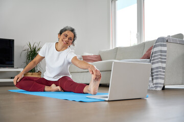 Happy middle aged older woman stretching leg at home looking at laptop. Fit sporty mature lady...