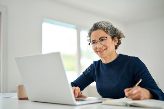 Happy middle aged woman sitting at table at home office using laptop learning online writing notes. Mature 50 years old lady wearing glasses remote working online on computer in living room.