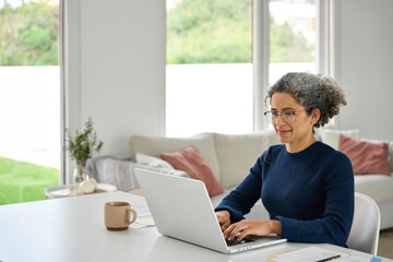 Happy middle aged woman sitting at table at home and using laptop in modern house. Mature 50 years...