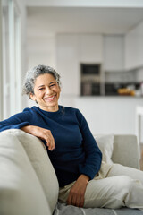 Happy middle aged woman sitting on comfy sofa in living room at home laughing. Smiling mature older...