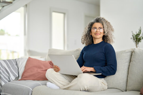 Happy curly middle aged woman using pc laptop surfing online sitting on sofa at home. Mature older lady looking away relaxing on couch with computer device in modern house living room.