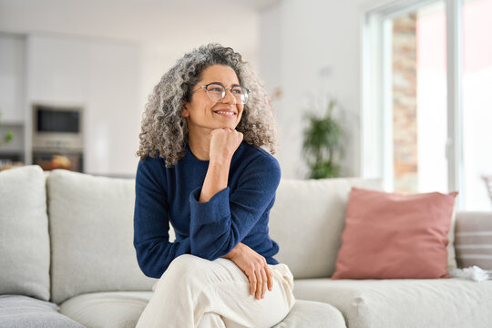 Happy relaxed 50 years old woman sitting on sofa at home looking away. Smiling middle aged lady posing in living room at home. Mature older female with curly gray hair relaxing on couch enjoying chill