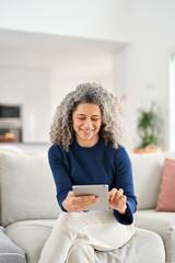 Smiling relaxed middle aged curly woman holding tab in hands relaxing on sofa at home. Happy mature...