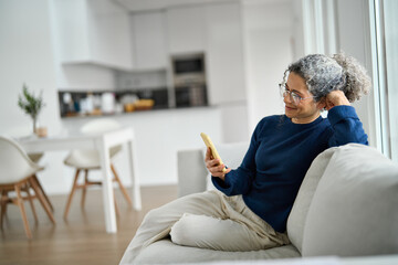 Happy middle aged woman using mobile phone messaging relaxing on couch at home. Smiling older...