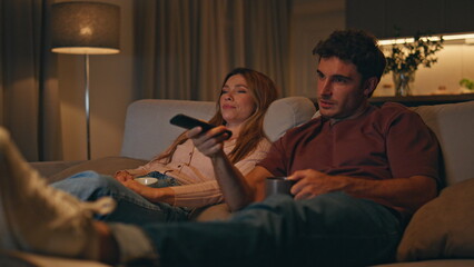 Fototapeta premium Smiling spouses watching tv sitting at cozy living room interior late evening