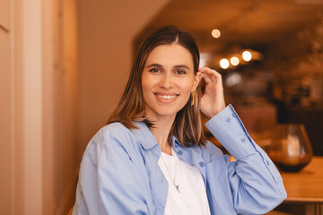 Cute brunette short hair girl smiling and fix hair. Woman fix her hair, look at camera. She wear blue shirt, white top, sitting rest in cafe. Happy brunette girl look flirting and coquettish.