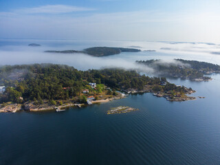 Morning fog rolling over an island in early autumn, in the Swedish archipelago, Sweden. Houses, trees in the mist. Blue sky, bright sunlight