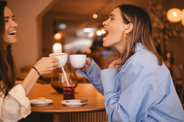 Communication and friendship concept - smiling young women sisters friends with coffee cups at cafe laughing and talking, they have good time together. Happy smiling girls gossiping.