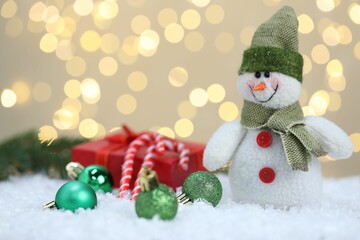 Funny snowman figure, Christmas balls, gift box and candy canes on artificial snow against blurred lights