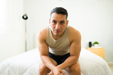 Strong young man looking at camera while sitting on bed and relaxing in his bedroom at home