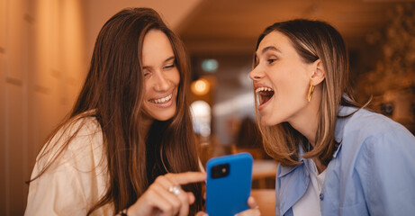 Portrait of excited impressed two  women sisters friends fellows use devices recommend select choose decide discount options, point finger on phone, screaming wow, open mouth, omg, sitting in cafe.