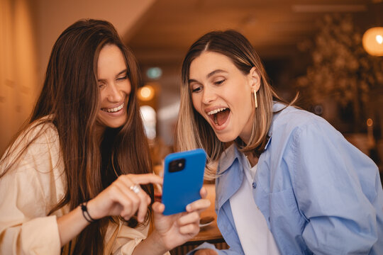 Portrait of excited impressed two women sisters friends fellows use devices recommend select choose decide discount options, point finger on phone, screaming wow, open mouth, omg, sitting in cafe.