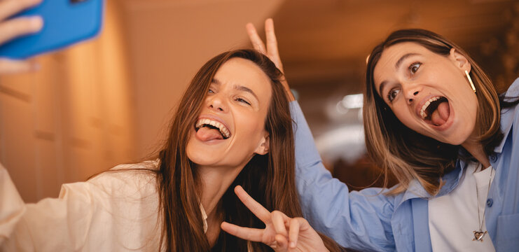 Two smiling hipster sisters friends female in trendy summer clothes. Carefree women rest sitting in cafe restaurant. Positive models having fun and taking selfie on smartphone, show peace v tongue out