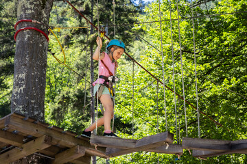 A girl in a pink shirt and blue helmet cautiously moves through a high ropes course in the woods, walking on a narrow wooden platform high above the ground, surrounded by lush green trees