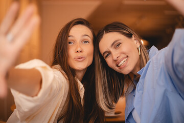 Two young beautiful smiling hipster sisters friends female in trendy summer clothes. Carefree women rest sitting in cafe restaurant. Positive models having fun and taking selfie on smartphone. 