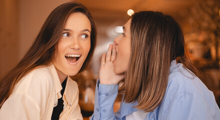 Gossiping. Two beautiful young women gossiping telling something on ear while sitting in the cafe. Communication and friendship concept. Young woman telling her friend sister some secrets. They cover.