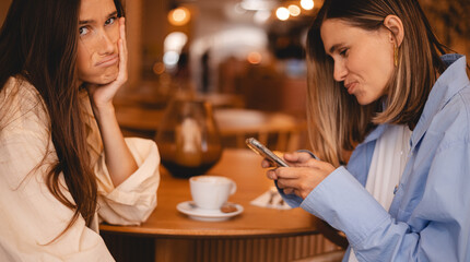 Upset girl sadly looking aside while another girl happy using cellphone in cozy of cafe. Unhappy woman sitting hold head, look boring while her sister chatting in mobile phone and smiling.