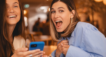 Beautiful amazing happy excited young two friends sisters women sitting in cafe hold mobile phone,...