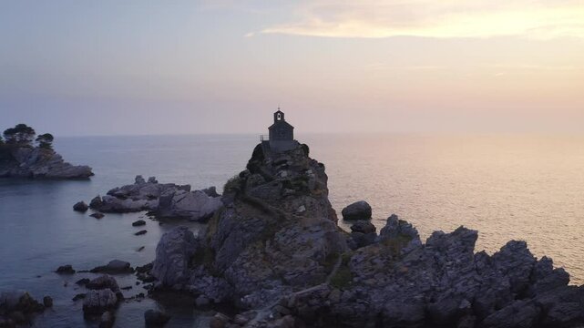 Flying drone 4K video around Sveta Nedjelja Island with magestic small church on cliff's top during evening sunset time hours. Balkans traveling, landmarks and Nature concept in Petrovac, Montenegro.