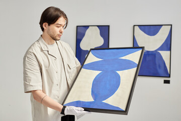 Minimal waist up portrait of young man as curator carefully holding abstract art painting while...