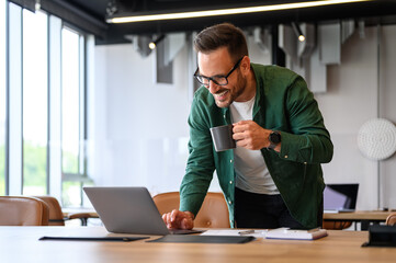 Smiling male freelancer drinking coffee and checking e-mails over wireless computer on desk in office