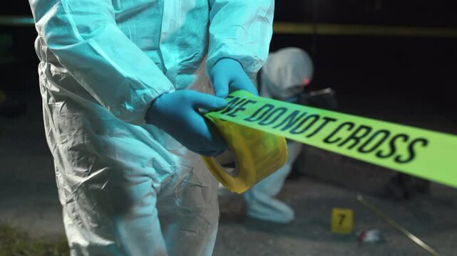 A forensic scientist rolling out police tape at a crime scene at night, while other experts collect evidence in the background.