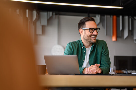 Happy young businessman in eyeglasses looking away while working online over laptop at desk in office