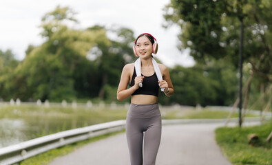 Invigorated Morning Run: Young Asian woman enjoys a scenic jog in the park, headphones in and towel...