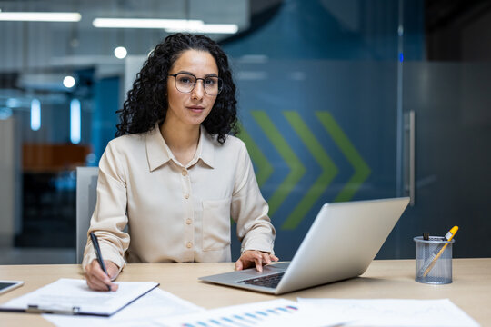Hispanic businesswoman deeply engaged in work at desk with laptop, pen in hand. Demonstrates focus and analytical thinking in modern office environment, reflecting professional dedication