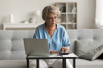 Focused mature woman using calculator, tallies up amounts due for utilities, credit cards, and...