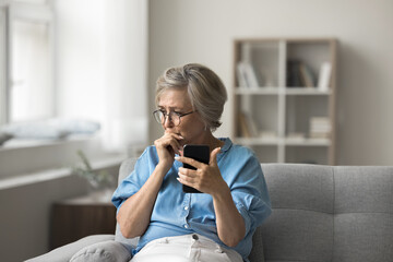 Unhappy older woman sitting on couch, holding smartphone feels frustrated and concerned, received upsetting news in sms, experiencing personal or family problems, misunderstanding through messages