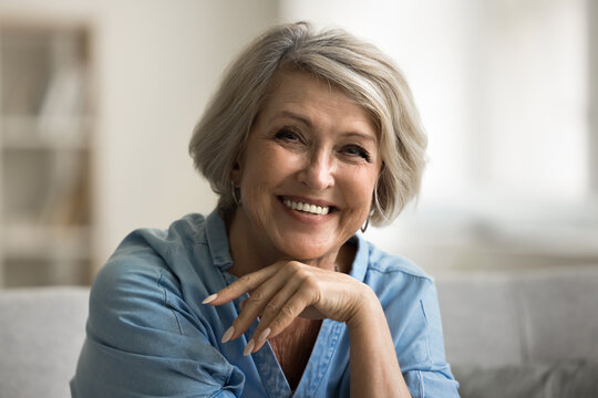 Head shot close up portrait beautiful mature woman posing for camera having perfect white-toothed smile sit alone on sofa at home, advertises professional dental services. Happy retirement and beauty