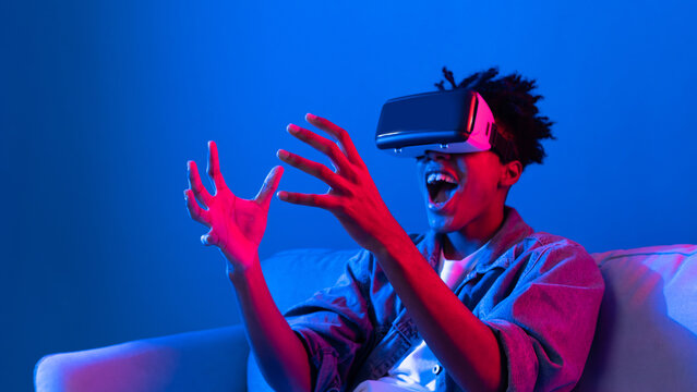Young African American looking VR enjoy watching fantastic 3D pointing interesting object technology blue neon light virtual reality metaverse new world at living room sitting on sofa. Contrivance.